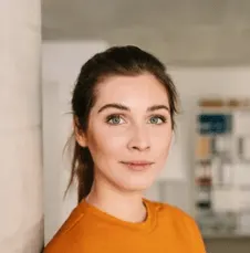 Woman with dark hair pulled back wearing an orange shirt, looking toward the camera in an indoor setting
