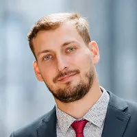 Muž s blond vlasy a vousem v tmavém saku a červené kravatě, portrét na neutrálním pozadí